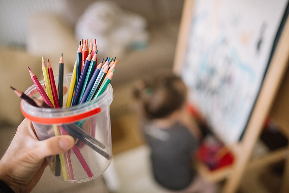 A hand holds a container filled with vibrant colored pencils. In the background