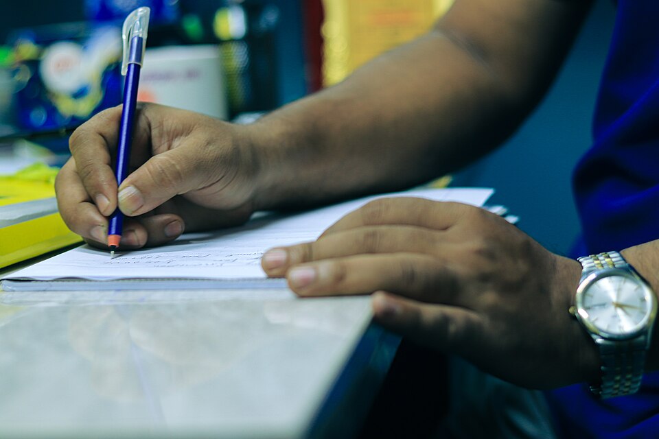 Person writing in Bengali script using a red ballpoint pen 04